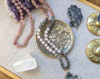 May include: A crystal mala necklace with pink and grey beads, a large iridescent labradorite pendant, and a small crystal cluster. The necklace is displayed on a decorative plate with a gold dragon design. There are also two small golden singing bowls and a clear crystal on the surface.