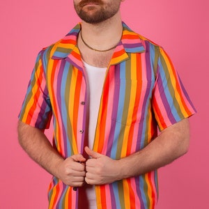 Pode incluir: Uma camisa de botões de manga curta com listras verticais em um padrão de cores do arco-íris. A camisa tem gola e um bolso do lado esquerdo.