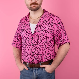 May include: A man wearing a pink short-sleeved shirt with a black geometric pattern, blue jeans, and a gold chain necklace. He is wearing glasses and has a shaved head.