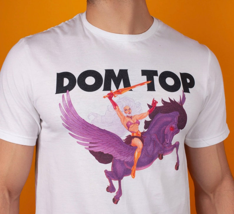 Pode incluir: Camiseta branca com uma estampa preta das palavras "DOM TOP" e um gr&aacute;fico de uma mulher montando um cavalo alado.