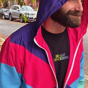Pu&ograve; includere: Giacca con cappuccio in stile retr&ograve;, con pannelli blu, rosa e viola. La giacca &egrave; chiusa con la zip, rivelando una maglietta nera con la scritta ricamata "BE GAY DO CRIMES".