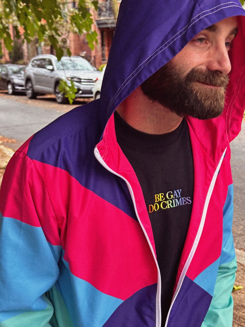 Pu&ograve; includere: Giacca con cappuccio in stile retr&ograve;, con pannelli blu, rosa e viola. La giacca &egrave; chiusa con la zip, rivelando una maglietta nera con la scritta ricamata "BE GAY DO CRIMES".