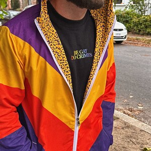 Pu&ograve; includere: Giacca con zip dai colori vivaci, con un design a blocchi di colore viola, giallo e rosso. La giacca ha un cappuccio e una cerniera bianca. L'interno del cappuccio presenta un motivo leopardato. &Egrave; visibile una maglietta nera con la scritta "BE GAY DO CRIMES".
