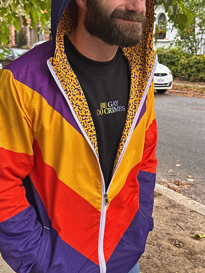 Pu&ograve; includere: Giacca con zip dai colori vivaci, con un design a blocchi di colore viola, giallo e rosso. La giacca ha un cappuccio e una cerniera bianca. L'interno del cappuccio presenta un motivo leopardato. &Egrave; visibile una maglietta nera con la scritta "BE GAY DO CRIMES".