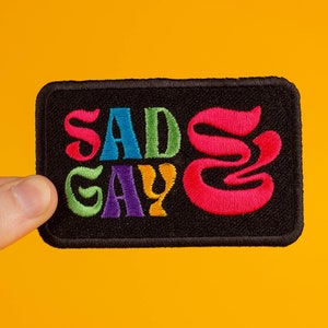 Könnte beinhalten: Ein schwarzer gestickter Aufnäher mit dem Text "Sad Gay" in Regenbogenfarben. Das "S" in "Sad" ist ein rosa Wirbel.