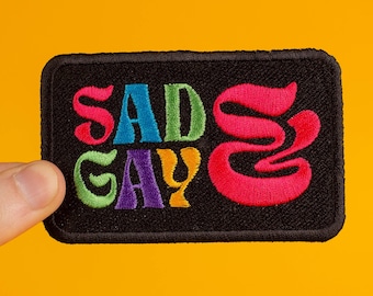 Puede incluir: Un parche bordado negro con el texto "Sad Gay" en colores arcoíris. La "S" de "Sad" es un remolino rosa.