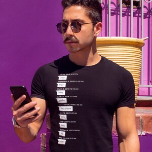 Op de afbeelding: Een persoon draagt een zwart T-shirt met witte tekst erop. De tekst vermeldt datums en tijden met het woord "Hey" of "Hello" naast elke datum. De persoon houdt een zwarte smartphone in zijn rechterhand.