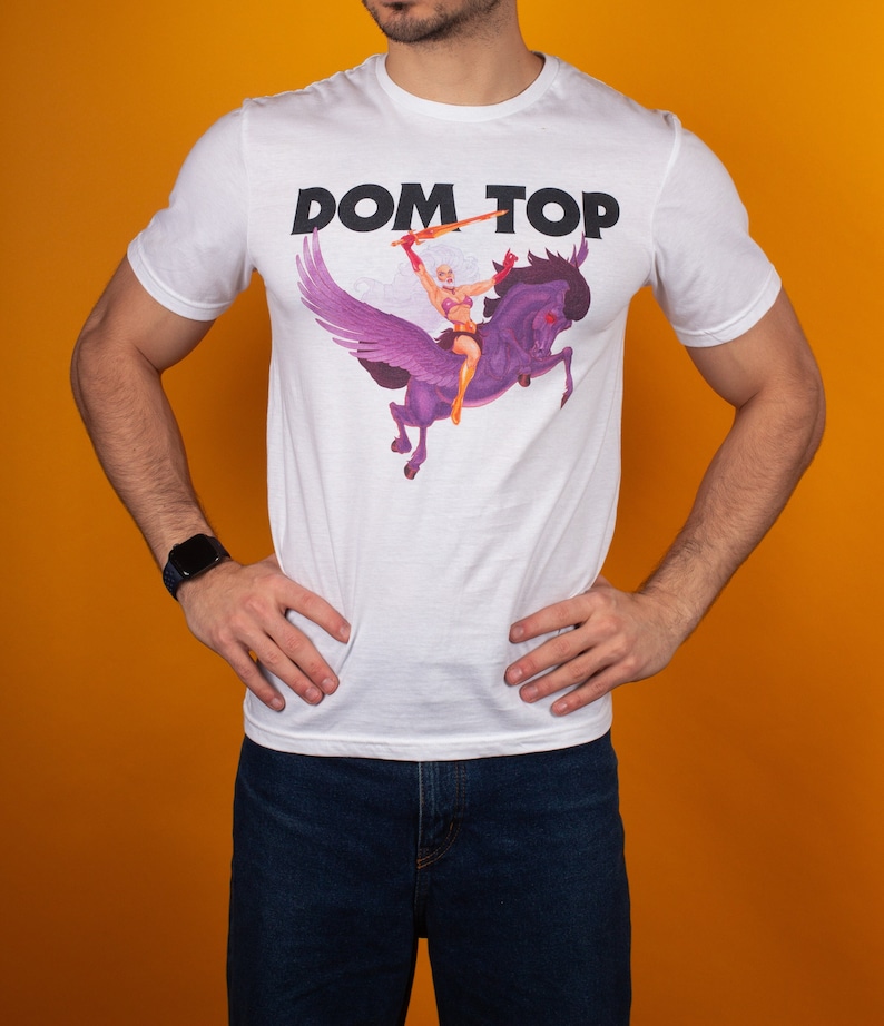 Pode incluir: Camiseta branca com uma estampa gr&aacute;fica de uma mulher montando um cavalo alado. O texto "DOM TOP" est&aacute; impresso em letras pretas acima do gr&aacute;fico.