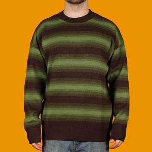 May include: A brown and green striped sweater. The sweater has horizontal stripes in varying shades of brown and green. The sweater has a crew neck and long sleeves. The background is a solid orange color.