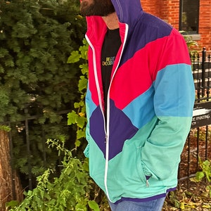 Pu&ograve; includere: Giacca con cappuccio multicolore con chiusura a zip. La giacca presenta un design a blocchi di colore con pannelli viola, rossi, turchese e verdi. La giacca &egrave; indossata sopra una maglietta nera con la scritta "BE GAY DO CRIM". La persona indossa jeans blu.