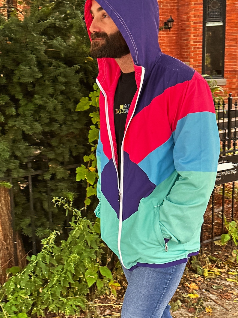 Pu&ograve; includere: Giacca con cappuccio multicolore con chiusura a zip. La giacca presenta un design a blocchi di colore con pannelli viola, rossi, turchese e verdi. La giacca &egrave; indossata sopra una maglietta nera con la scritta "BE GAY DO CRIM". La persona indossa jeans blu.
