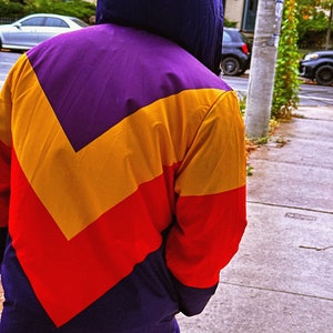 Pu&ograve; includere: Una giacca con cappuccio con un design a blocchi di colore. La giacca presenta un cappuccio viola e un motivo a chevron giallo, rosso e viola. La parte inferiore della giacca &egrave; di colore viola scuro.