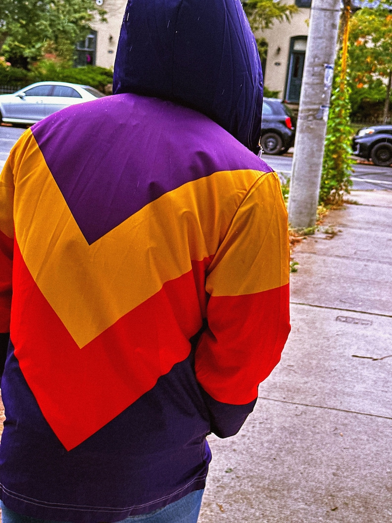 Pu&ograve; includere: Una giacca con cappuccio con un design a blocchi di colore. La giacca presenta un cappuccio viola e un motivo a chevron giallo, rosso e viola. La parte inferiore della giacca &egrave; di colore viola scuro.