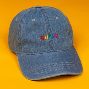 May include: A blue denim baseball cap with the word "QUEER" embroidered in rainbow colours.