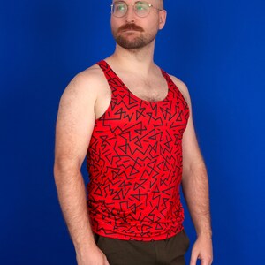 以下が含まれることがあります： 青い背景の前に立っている男性。赤いタンクトップを着ており、黒い幾何学模様が描かれている。茶色のズボンを履いている。