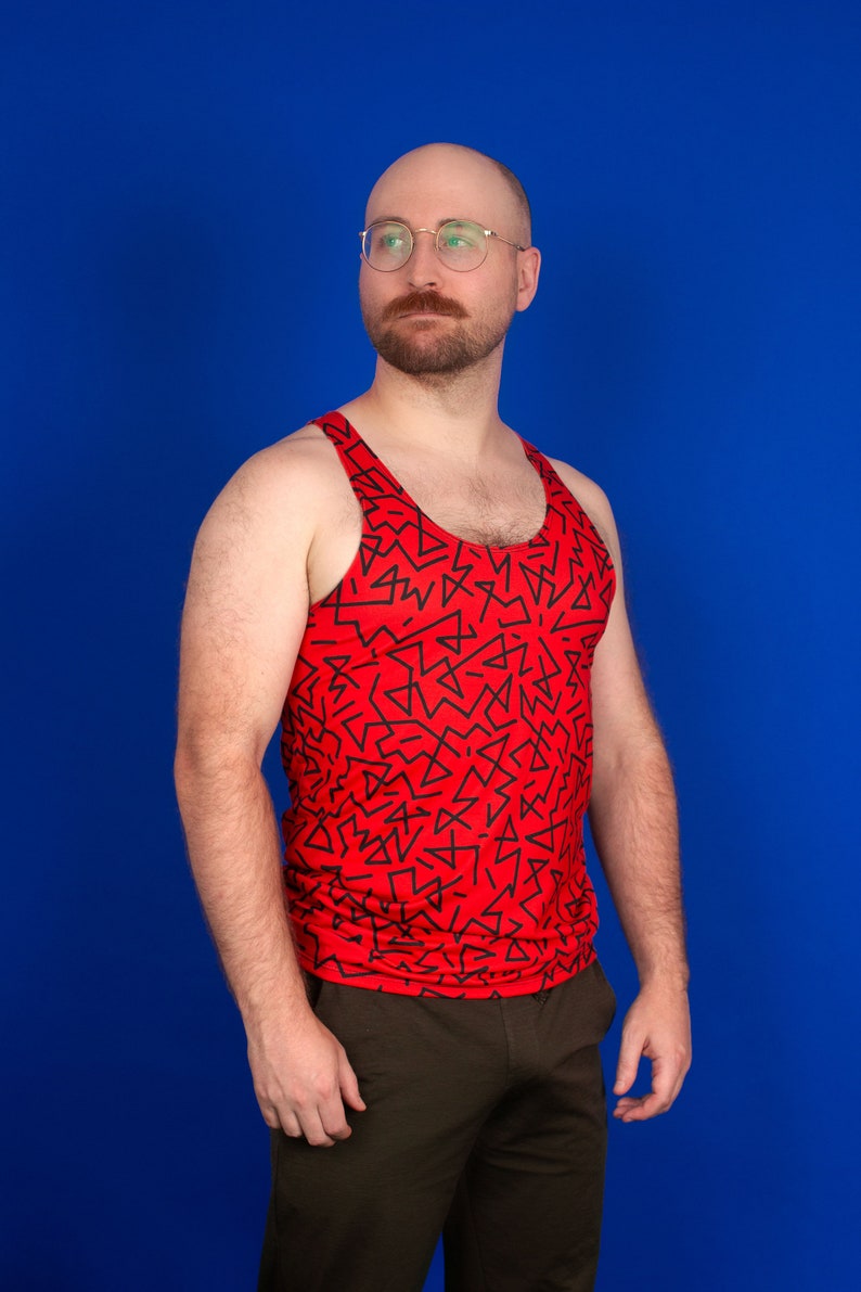 以下が含まれることがあります： 青い背景の前に立っている男性。赤いタンクトップを着ており、黒い幾何学模様が描かれている。茶色のズボンを履いている。