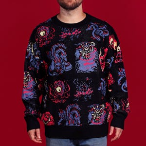 Puede incluir: Jersey negro de cuello redondo con un diseño de monstruos pixelados repetidos en rojo, morado y azul. El jersey tiene puños negros y cuello negro. El fondo es rojo liso.