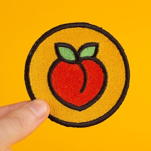 May include: A yellow and black embroidered patch with a red peach design. The peach has a green stem and leaves.