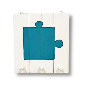 Könnte beinhalten: Weißer Holz-Schlüsselhalter mit drei Haken. Der Schlüsselhalter ist in Form eines Puzzlesteins geformt und in Türkis lackiert.