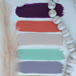 Puede incluir: Un conjunto de seis muestras de pintura en varios colores, incluyendo morado, naranja, beige, verde, gris y amarillo. Las muestras están dispuestas verticalmente sobre un fondo blanco. Una hebra de cuentas de madera blancas con un borla marrón está en el lado derecho de la imagen.