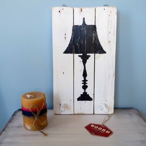 May include: A white wooden sign with a black silhouette of a table lamp. The sign has two white knobs on the bottom and is hanging from a hook. A candle and a red tag with the text "WOORD" are on a white wooden surface.