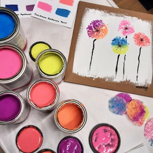 May include: A collection of paint pots in various colors, including pink, purple, orange, yellow, and blue. The paint pots are arranged on a white surface, along with a cardboard sheet with a painting of colorful dandelions. The text "Daydream Apothecary Paint" is visible on the cardboard sheet.