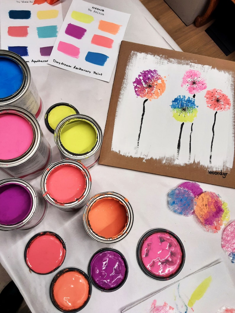 May include: A collection of paint pots in various colors, including pink, purple, orange, yellow, and blue. The paint pots are arranged on a white surface, along with a cardboard sheet with a painting of colorful dandelions. The text "Daydream Apothecary Paint" is visible on the cardboard sheet.