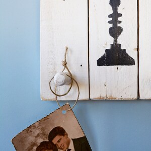 May include: A vintage photograph of a couple, a woman with short brown hair and a man with short brown hair, is hanging from a white wooden hook with twine. The couple is smiling and holding a bouquet of flowers. The hook is attached to a white wooden wall with a black painted design.