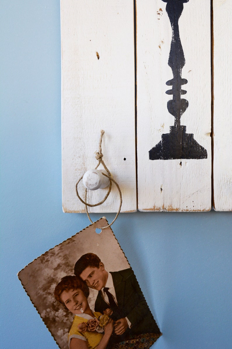 May include: A vintage photograph of a couple, a woman with short brown hair and a man with short brown hair, is hanging from a white wooden hook with twine. The couple is smiling and holding a bouquet of flowers. The hook is attached to a white wooden wall with a black painted design.