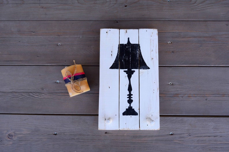 May include: A white painted wooden sign with a black silhouette of a table lamp. The sign has two hooks on the bottom. A small, wrapped candle is on the left side of the sign.