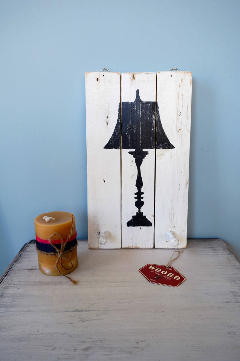 May include: A white wooden sign with a black silhouette of a table lamp. The sign has two white knobs on the bottom and is hanging from a hook. A candle and a red tag with the text "WOORD" are on a white wooden surface.