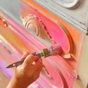 May include: A hand holding a paintbrush, painting a pink drawer with a decorative detail. The drawer is part of a dresser with a multi-colored ombre finish.