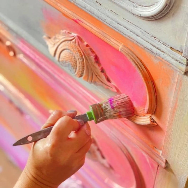 May include: A hand holding a paintbrush, painting a pink drawer with a decorative detail. The drawer is part of a dresser with a multi-colored ombre finish.