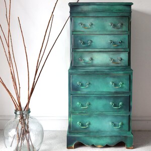 Puede incluir: Una c&oacute;moda alta de estilo antiguo con cinco cajones. La c&oacute;moda est&aacute; pintada en un acabado verde azulado desgastado con herrajes dorados. La c&oacute;moda tiene una base ligeramente curvada y se encuentra sobre una superficie blanca.