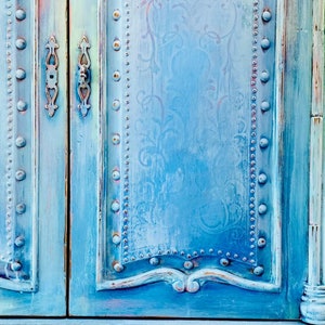 May include: A distressed blue cabinet with two doors and decorative metal accents. The cabinet has a light blue and white floral pattern on the front.