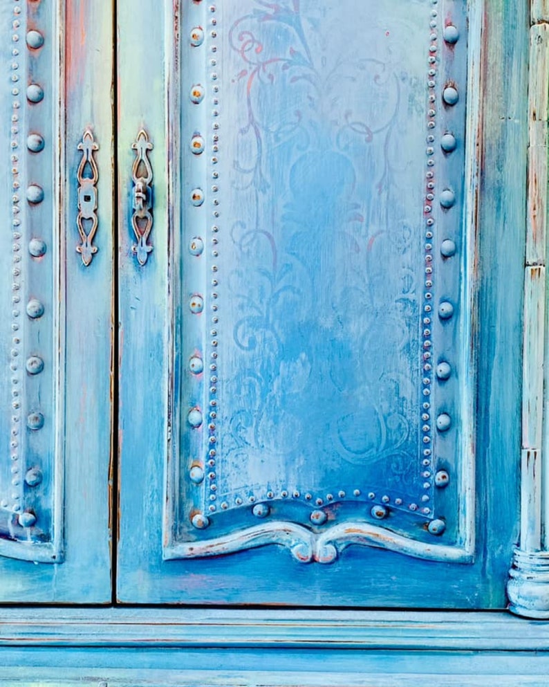 May include: A distressed blue cabinet with two doors and decorative metal accents. The cabinet has a light blue and white floral pattern on the front.