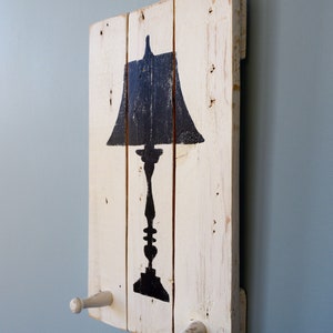 May include: A white painted wooden sign with a black silhouette of a lamp. The sign has two white wooden hooks on the bottom.
