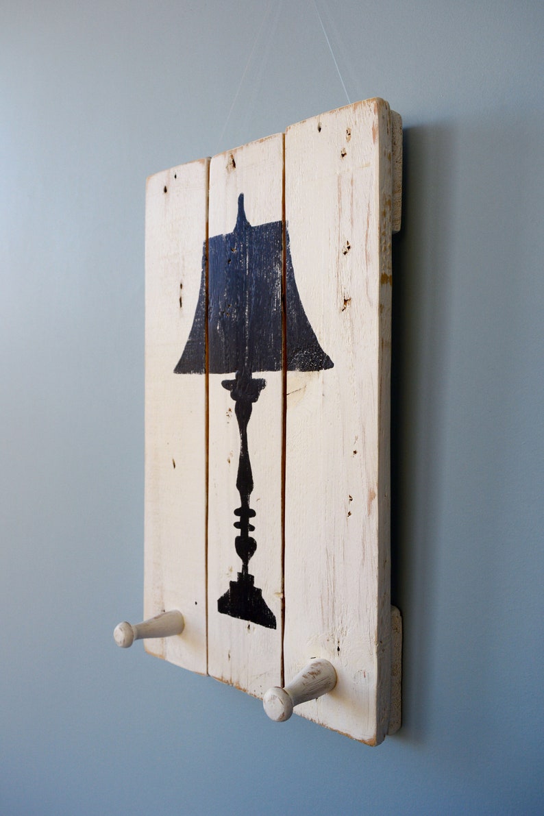 May include: A white painted wooden sign with a black silhouette of a lamp. The sign has two white wooden hooks on the bottom.