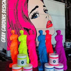 May include: Six neon-colored figurines of the Venus de Milo statue, with a pop art style image of a woman's face in the background.  The figurines are arranged in a row, with the colors ranging from orange to purple.  There are six jars of paint in the foreground, each with a different color of paint to match the figurines.  The paint jars are labeled "Gray Gardens Design."