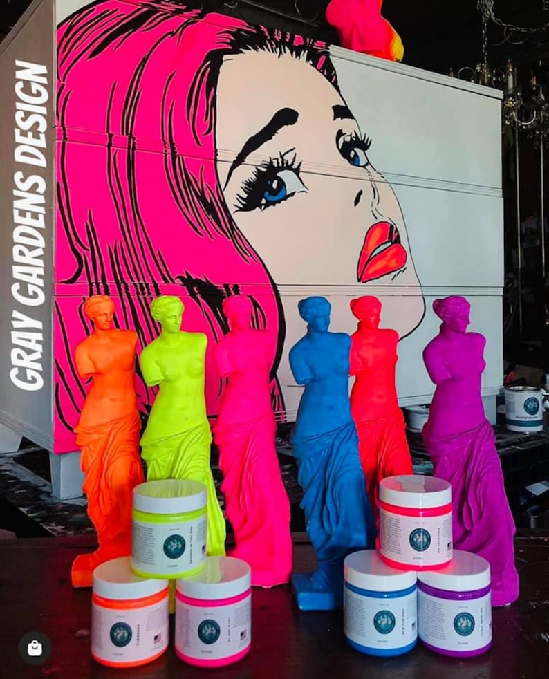 May include: Six neon-colored figurines of the Venus de Milo statue, with a pop art style image of a woman's face in the background.  The figurines are arranged in a row, with the colors ranging from orange to purple.  There are six jars of paint in the foreground, each with a different color of paint to match the figurines.  The paint jars are labeled "Gray Gardens Design."