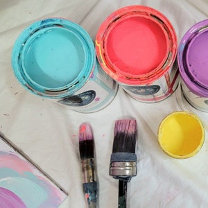 May include: Three paint cans with lids, one blue, one pink, and one purple, with two paint brushes on a white surface.