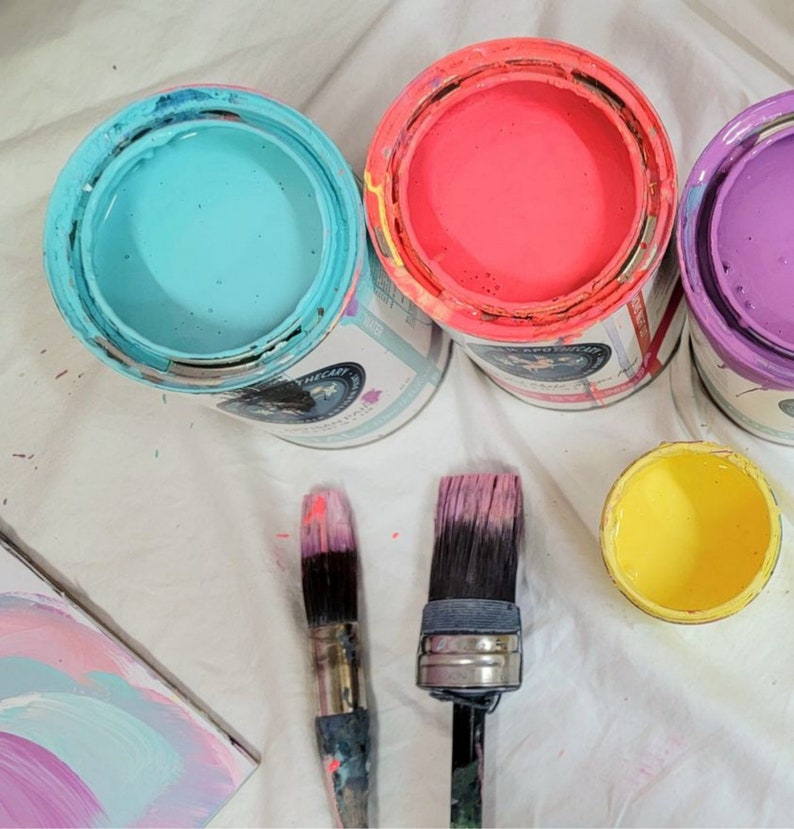 May include: Three paint cans with lids, one blue, one pink, and one purple, with two paint brushes on a white surface.