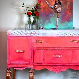 May include: A vintage pink painted buffet with gold hardware and a white distressed top. The buffet has four drawers and two doors. The buffet is sitting on a white floor.