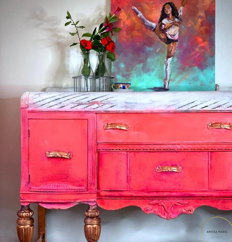 May include: A vintage pink painted buffet with gold hardware and a white distressed top. The buffet has four drawers and two doors. The buffet is sitting on a white floor.