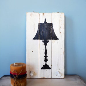 May include: A whitewashed wooden sign with a black silhouette of a table lamp. The sign has two white knobs on the bottom. A brown candle is in the foreground.
