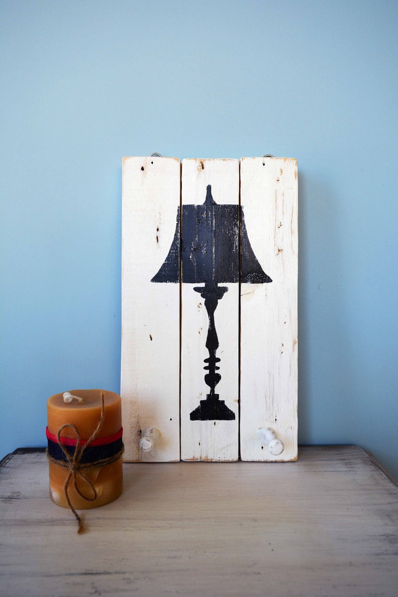 May include: A whitewashed wooden sign with a black silhouette of a table lamp. The sign has two white knobs on the bottom. A brown candle is in the foreground.
