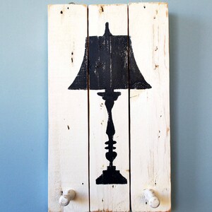 May include: White wooden wall decor with a black silhouette of a table lamp with a shade. The decor has two white wooden knobs on the bottom.