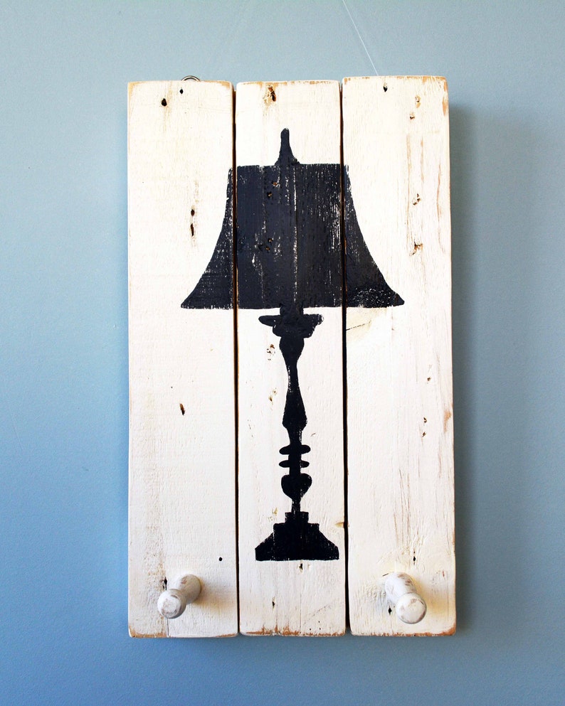 May include: White wooden wall decor with a black silhouette of a table lamp with a shade. The decor has two white wooden knobs on the bottom.