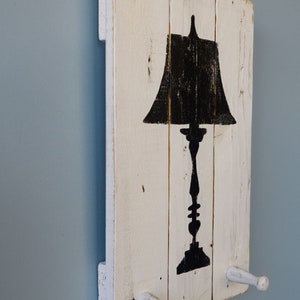 May include: White wooden wall decor with a black silhouette of a table lamp. The wood has a distressed look and there are two white wooden hooks on the bottom.