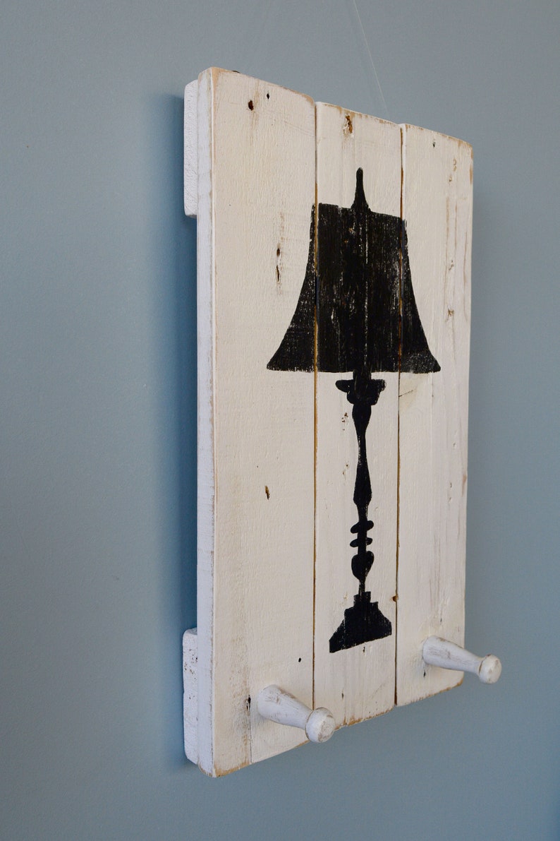 May include: White wooden wall decor with a black silhouette of a table lamp. The wood has a distressed look and there are two white wooden hooks on the bottom.